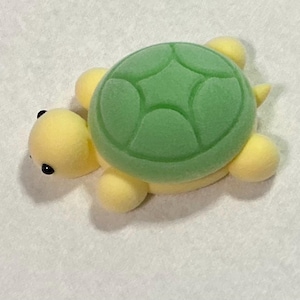 May include: A small, yellow and green turtle figurine. The turtle has a green shell with a textured pattern, yellow head, feet, and tail. The turtle has small black eyes.