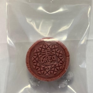 Op de afbeelding: Een rode Oreo-koekje met het woord "OREO" erop gedrukt, in een doorzichtige plastic zak. Twee kleine, zilverkleurige, fuzzy objecten bevinden zich onder in de zak.