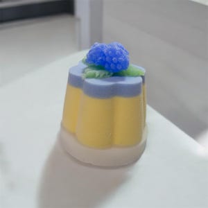 May include: A decorative, layered candle in the shape of a flower. The candle has a yellow base, a lavender middle layer, and a blue floral top. Green leaves accent the blue flower. The candle sits on a white base.
