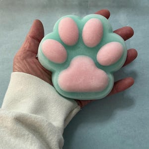 Puede incluir: Un jabón en forma de pata azul claro con almohadillas rosas. El jabón se sostiene en una mano, mostrando los colores pastel y el diseño detallado de la huella de la pata. Es un artículo único y decorativo.