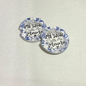 May include: Two white car coasters with a floral design in shades of blue and gray. The coasters have the text "My Squad Calls Me Mama" in black script and block letters.