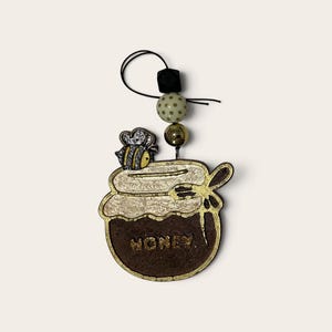 May include: A decorative hanging ornament shaped like a honey jar with a bee on the lid and the word HONEY printed on the front. The jar is brown with a gold rim and features a small wooden dipper. It hangs from a black string with three beads.