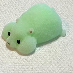 May include: A light green, plush hippo-shaped toy with black eyes and small, light brown ears. The toy has a rounded body and is resting on a white, textured surface. The hippo has a small tail.