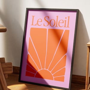 May include: Framed art print with the words "Le Soleil" above an orange sunburst design on a pink background. The artwork is in a black frame and leans against a white wall.