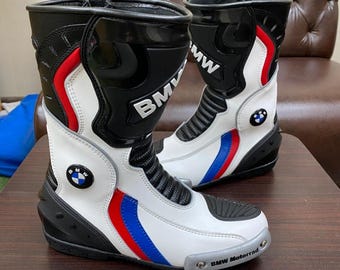 Puede incluir: Par de botas de moto blancas y negras con rayas rojas y azules. Las botas tienen el logotipo de BMW y la inscripción "BMW Motorrad". Fabricadas en cuero, tienen una suela de goma.