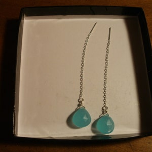 May include: A pair of silver dangle earrings with turquoise blue gemstone drops. The earrings are displayed in a black box.