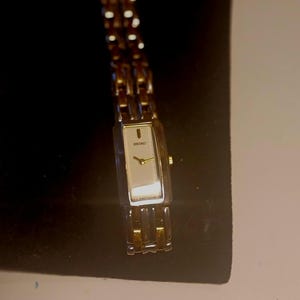 May include: A rectangular Seiko watch with a gold-toned band and face. The watch has a white face with gold-toned hands and markers. The band is made of small, connected links. The watch is displayed on a black surface.