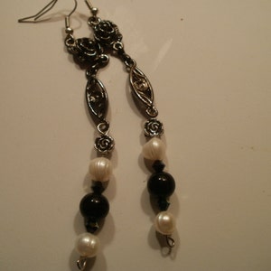 May include: A pair of long silver earrings with black and white beads. The earrings feature a floral design and are accented with black beads.