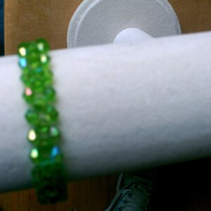 May include: A green crystal beaded bracelet with iridescent facets. The bracelet is displayed on a white mannequin arm.