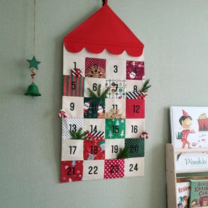 Może przedstawiać: Materiałowy kalendarz adwentowy z czerwonym dachem i 24 ponumerowanymi kieszeniami. Kieszenie zdobione są świątecznymi wzorami, a niektóre zawierają małe gałązki zieleni i laski cukrowe. Zielona gwiazda i dzwonek wiszą po lewej stronie.
