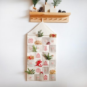 Pode incluir: Um calendário do advento de Natal em tecido com 24 bolsos numerados. Os bolsos apresentam um padrão xadrez bege e branco, com desenhos natalinos bordados em vermelho. Pequenos ramos de sempre-vivas e presentes em miniatura são colocados em alguns bolsos.