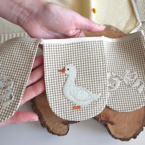 May include: A handmade, beige and white gingham banner with appliqued duck and bow designs. The banner is held up by a person's hand, and is resting on a wooden surface. The banner is part of a baby or nursery decor.