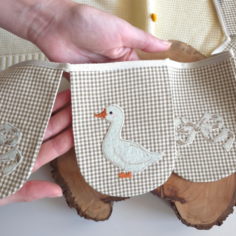 May include: A handmade, beige and white gingham banner with appliqued duck and bow designs. The banner is held up by a person's hand, and is resting on a wooden surface. The banner is part of a baby or nursery decor.