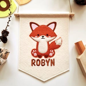 Könnte beinhalten: Ein cremefarbenes Filzbanner mit einem gestickten Cartoon-Fuchs in Orange, Braun und Weiß. Der Fuchs sitzt mit ausgestreckten Pfoten. Der Name "ROBYN" ist in braunen Buchstaben darunter gestickt. Das Banner hängt an einer Holzstange.