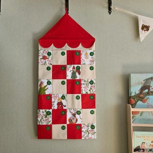 Peut inclure: Un calendrier de l'Avent en tissu à suspendre avec un sommet pointu rouge à bord festonné. Il présente une grille de vingt-quatre poches carrées en tissu rouge, crème et à motifs, numérotées de un à vingt-quatre dans des cercles verts.