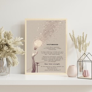 May include: A framed art print with the word "VICTIMHOOD" and text about inner strength. The artwork features a lightbulb, a hand reaching up, and a power pole illustration. The colour palette is neutral with beige and brown tones.
