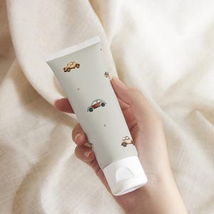 May include: A light gray tube with a white cap, decorated with small, colorful car illustrations. The tube is held in a hand and appears to be a skincare product, possibly a lotion or cream. The design gives a playful, vintage feel.