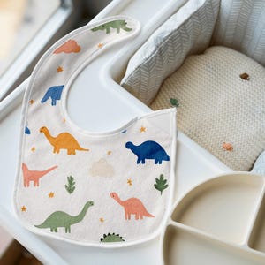 May include: A baby bib with a dinosaur pattern in orange, blue, green, and pink. The bib is on a white high chair tray, next to a divided plate and a cream-colored pillow.