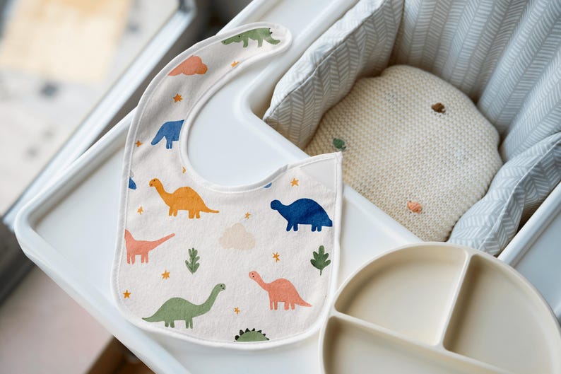 May include: A baby bib with a dinosaur pattern in orange, blue, green, and pink. The bib is on a white high chair tray, next to a divided plate and a cream-colored pillow.