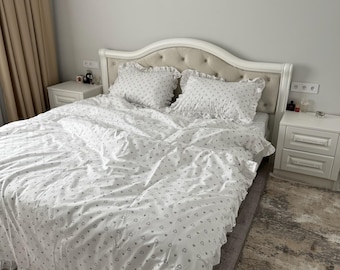May include: A white bedding set featuring a repeating pattern of small gray hearts. The set includes a duvet cover and two matching rectangular pillows, all finished with ruffled edges. The bedding is displayed on a tufted headboard bed frame.