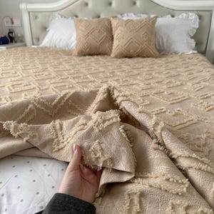Könnte beinhalten: Beige gesteppte Tagesdecke mit geometrischem Muster, ergänzt durch passende Zierkissen. Das Bett ist mit weißen Rüschenkissen und einem weißen Kopfteil ausgestattet. Eine Person hält die Tagesdecke.