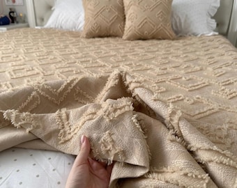 May include: A beige textured throw blanket with a raised geometric pattern and frayed edges, draped over a bed with white polka dot sheets and two matching beige square throw pillows against a tufted white headboard.