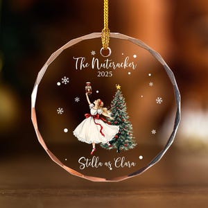 May include: A clear, circular Christmas bauble with a gold hanging loop. It features a ballerina illustration from "The Nutcracker" ballet, with the text "The Nutcracker 2025" and "Stella as Clara". Snowflakes and a Christmas tree are also depicted.