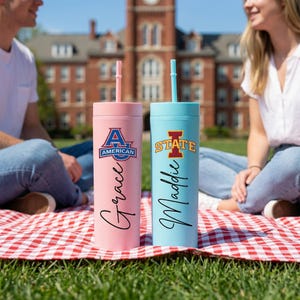 May include: Two insulated tumblers with straws. One is pink with "Grace" and "American" text, the other is light blue with "Maddie" and "STATE" text. Both tumblers have a matching straw and are on a red and white checkered blanket.