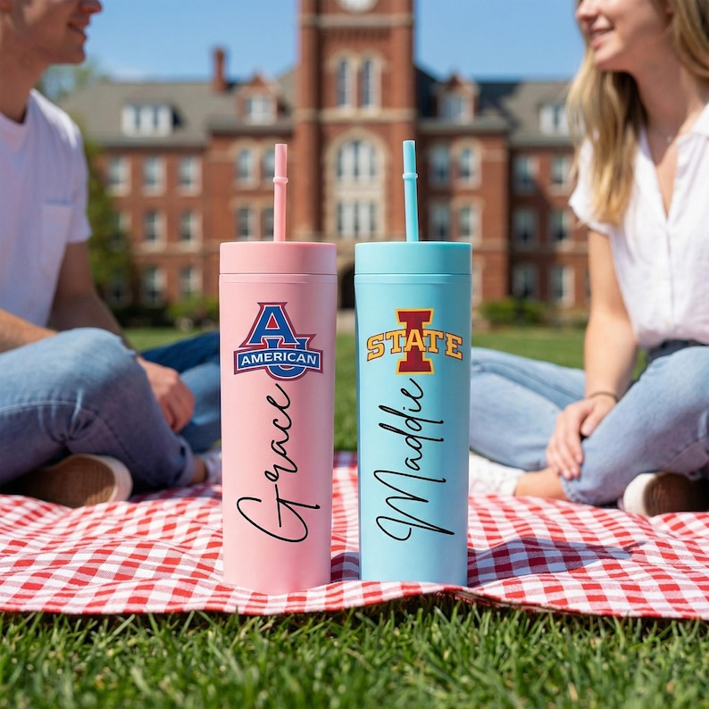 May include: Two insulated tumblers with straws. One is pink with "Grace" and "American" text, the other is light blue with "Maddie" and "STATE" text. Both tumblers have a matching straw and are on a red and white checkered blanket.