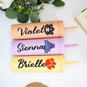May include: Three insulated tumblers in peach, lavender, and yellow. Each tumbler has a name in black script: Violet, Sienna, and Brielle. The Violet tumbler has a navy and orange logo. The Sienna tumbler has a blue and red logo. The Brielle tumbler has an orange paw print.
