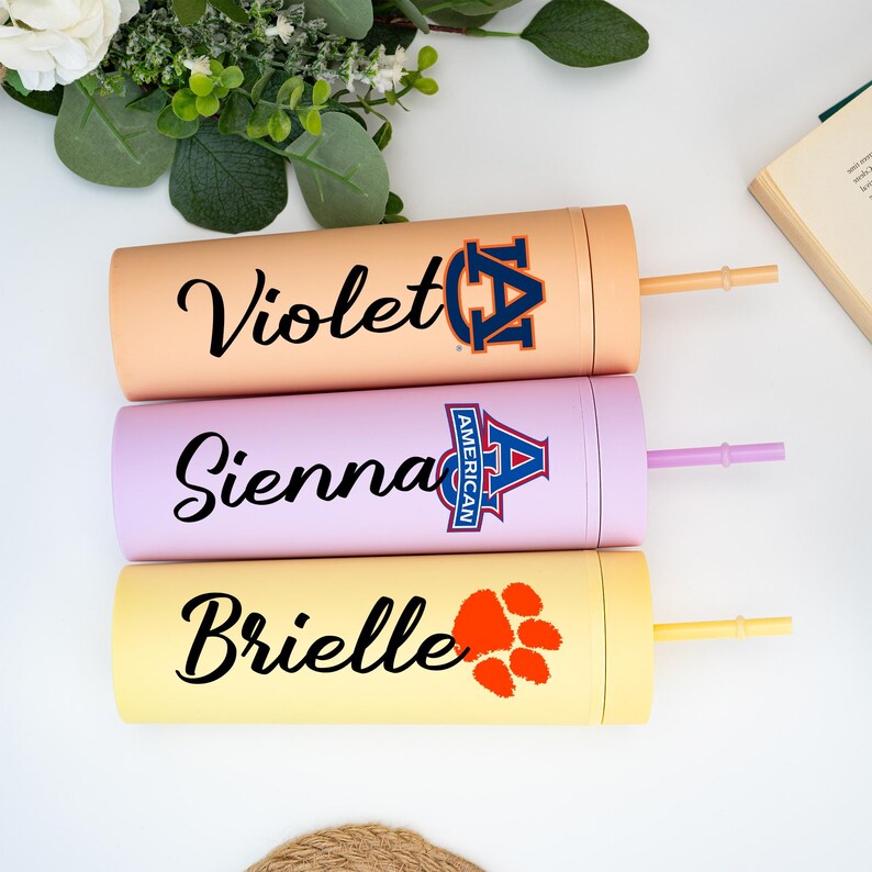 May include: Three insulated tumblers in peach, lavender, and yellow. Each tumbler has a name in black script: Violet, Sienna, and Brielle. The Violet tumbler has a navy and orange logo. The Sienna tumbler has a blue and red logo. The Brielle tumbler has an orange paw print.