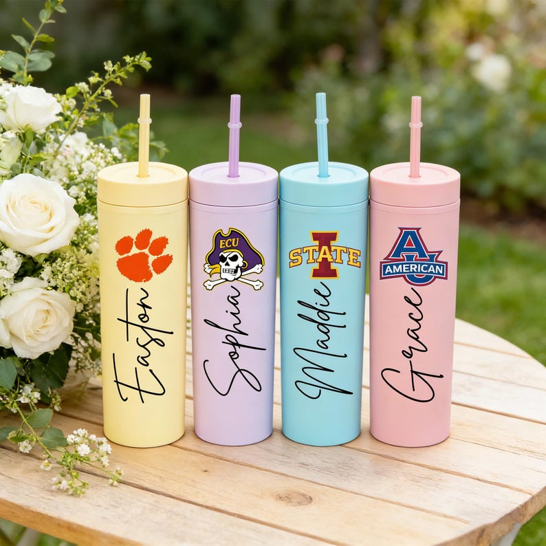 May include: Four pastel-coloured tumblers: yellow, lilac, light blue, and pink. Each tumbler has a different design, including a paw print, a skull and crossbones, a university logo, and a school logo, with a name in script.