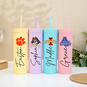 May include: Four pastel-coloured tumblers with lids and straws. Each tumbler is a different colour: yellow, lavender, light blue, and pink. Each has a different design and name printed on it.