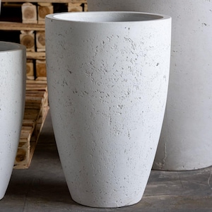 May include: A large, light grey concrete planter with a textured finish. The planter has a wide top and a narrowing base. Other similar planters are visible. Suitable for gardens or interiors.