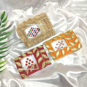 May include: Three rectangular bindi packs with TraditionalRoots21 text. One pack features gold fringe and multicolored bindis. The other two packs are red and orange with gold leaf embroidery and matching red bindis in the center.