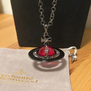 Könnte beinhalten: Eine silberfarbene Halskette mit einem Anhänger mit rotem Orb-Design, umgeben von einem schwarzen Ring. Der Anhänger ist mit Kristallen und einer kreuzförmigen Verzierung versehen. Ein kleiner Silberring und ein graues Samttuch sind ebenfalls sichtbar.