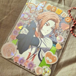 May include: A clear plastic card holder with a colorful anime illustration of a person with orange hair. The holder is decorated with stickers, including a butterfly, flowers, and hearts. The word "SENDO" is spelled out in orange letters.