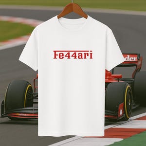 May include: White t-shirt with the red text "Fe44ari" on the front. The shirt hangs on a wooden hanger. A red and black race car is visible in the background.