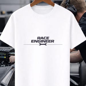 Puede incluir: Camiseta blanca con la frase "RACE ENGINEER" en azul oscuro y un gráfico de llave inglesa. La camiseta es de cuello redondo y parece estar hecha de un material suave.