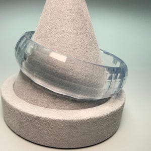 May include: A clear, light blue bangle bracelet with a faceted design. The bracelet is displayed on a gray, textured jewelry stand. The bangle's geometric pattern catches the light, creating a sparkling effect.