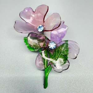 May include: A floral brooch featuring two translucent light purple petals with smooth, rounded edges. Each flower center is accented with a small, round, light blue rhinestone. The stem and leaves are painted in a vibrant, glossy green finish.