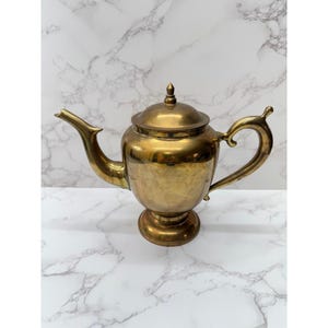 May include: A vintage brass teapot with a lid, spout, and curved handle. The teapot has a pedestal base and a shiny, golden finish. The background is a white marble pattern.