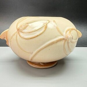 May include: A cream-colored ceramic bowl with a rounded, oval shape and a small pedestal base. The exterior features a raised, embossed floral pattern with light brown accents highlighting the petals and stems on the smooth, glossy surface.