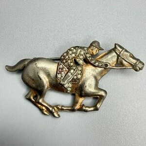 May include: A silver-toned metal brooch depicting a jockey riding a galloping horse. The jockey wears a cap and a suit embellished with small, clear, and multi-colored rhinestones. The horse is shown in a running pose with a bridle and reins.