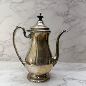 May include: A silver-colored coffee pot with a curved spout and handle. The pot has a lid with a black top and sits on a small, rounded base. The pot is reflective, showing the surrounding environment.