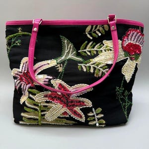 May include: Black tote bag with pink trim and handles, adorned with floral embroidery and sequin details. The bag features a vibrant design with red, pink, and cream-colored flowers and green foliage. The handles are a bright pink color.