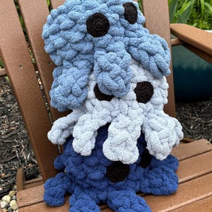 May include: Three handmade, plush octopus toys in shades of blue and gray, with black circular eyes. The toys are stacked on a wooden chair, showcasing their soft, textured appearance. A whimsical and cuddly item.