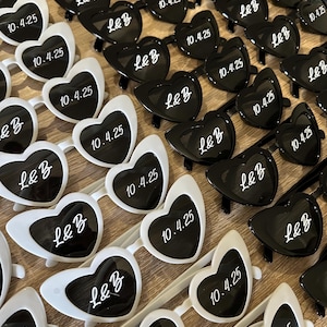 Può includere: Occhiali da sole a forma di cuore in bianco e nero, disposti in file. Le lenti presentano la scritta "L&B" e la data "10.4.25" in bianco. Un accessorio divertente per eventi.