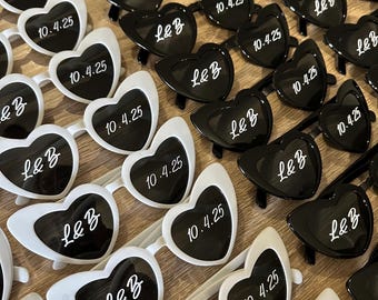 Puede incluir: Gafas de sol en forma de corazón en blanco y negro, dispuestas en filas. Las lentes muestran las letras "L&B" y la fecha "10.4.25" en blanco. Un accesorio divertido para eventos.