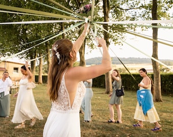 May include: A maypole featuring long, flowing ribbons in shades of sage green, cream, and white, radiating from a central point decorated with a floral arrangement, held by individuals in an outdoor setting with trees and a grassy field.